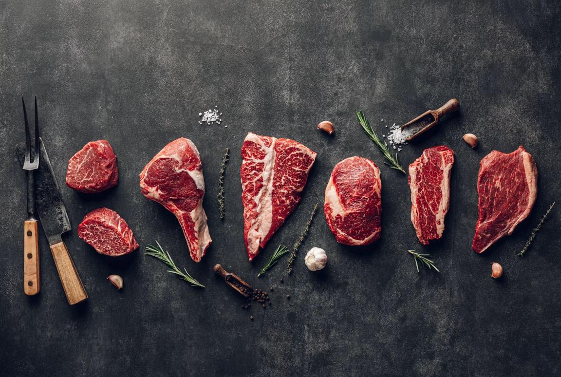 Raw beef cuts laid out on a dark slate counter with a carving fork, knife, rosemary and garlic